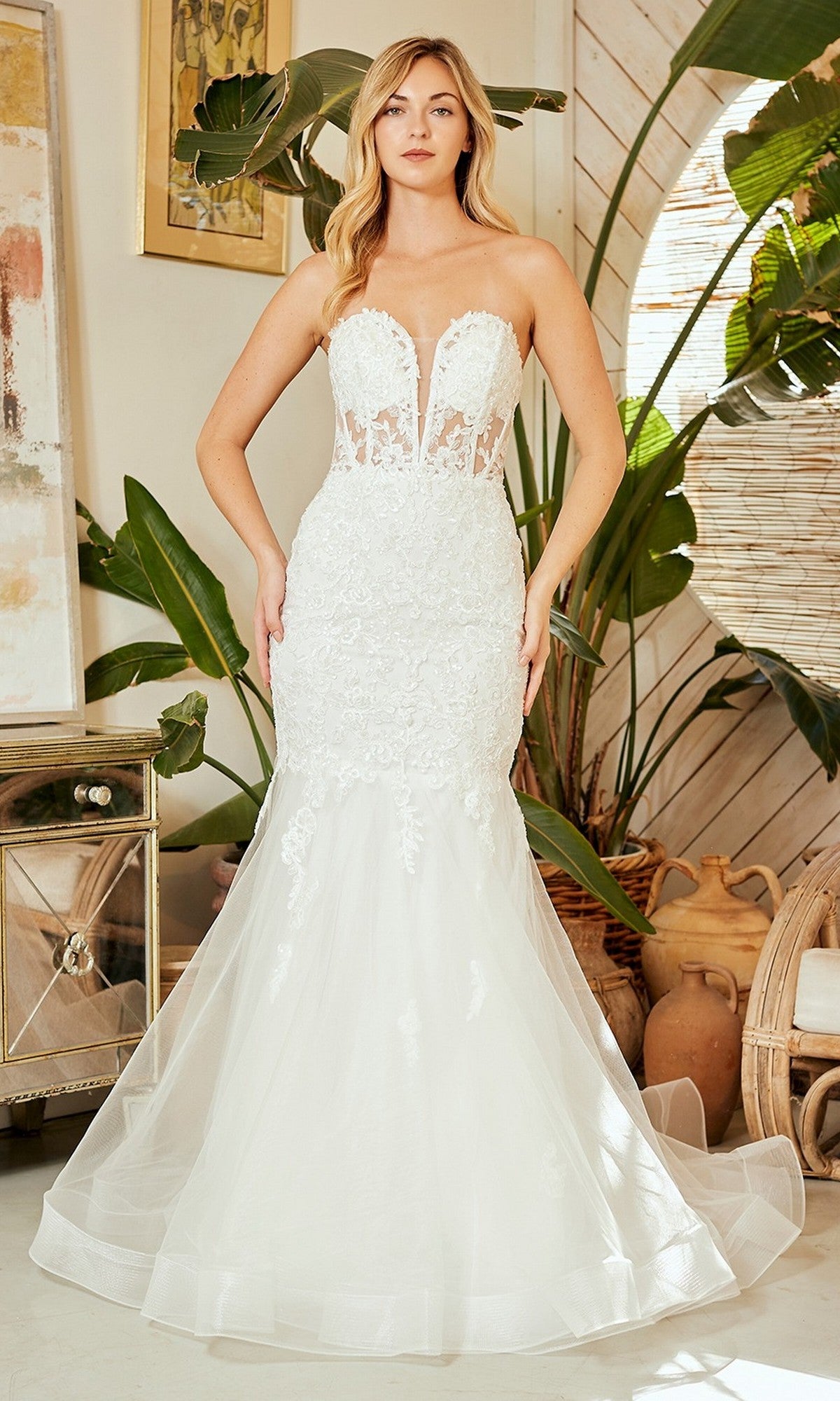 A woman stands indoors surrounded by plants and pottery, wearing a white Long Prom Dress: Amelia Couture 7090 featuring a strapless mermaid silhouette, lace details, sheer bodice, and tulle skirt.